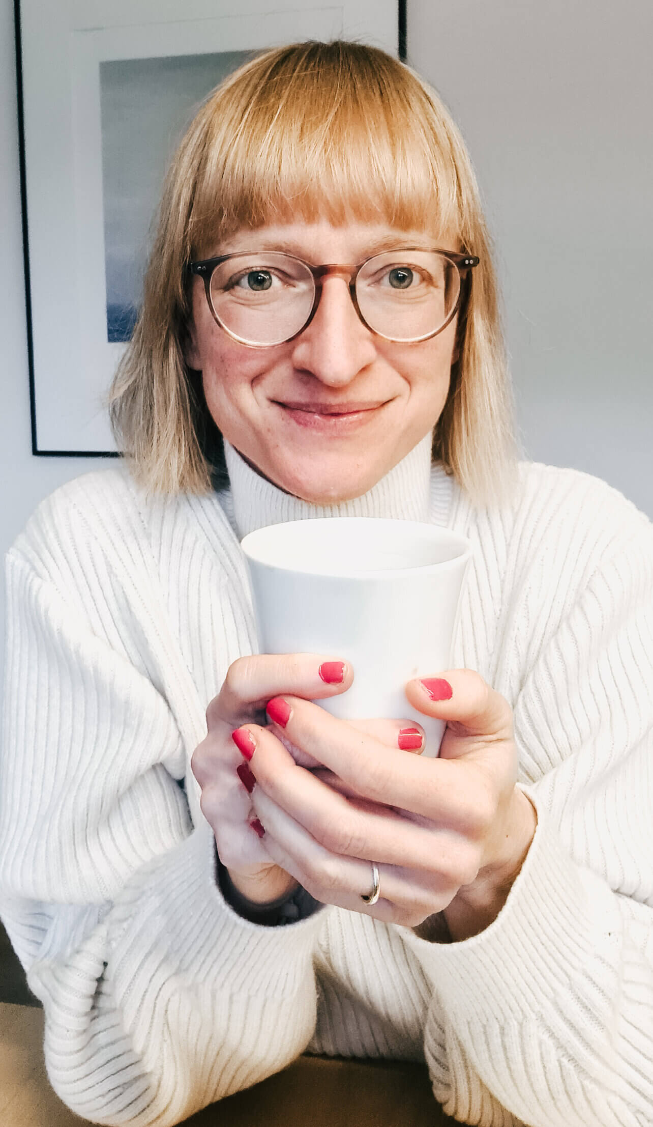 Marlis Schorcht sitzt am Tisch und schaut in die Kamera. SIe hält eine Tasse Kaffee in den Händen.