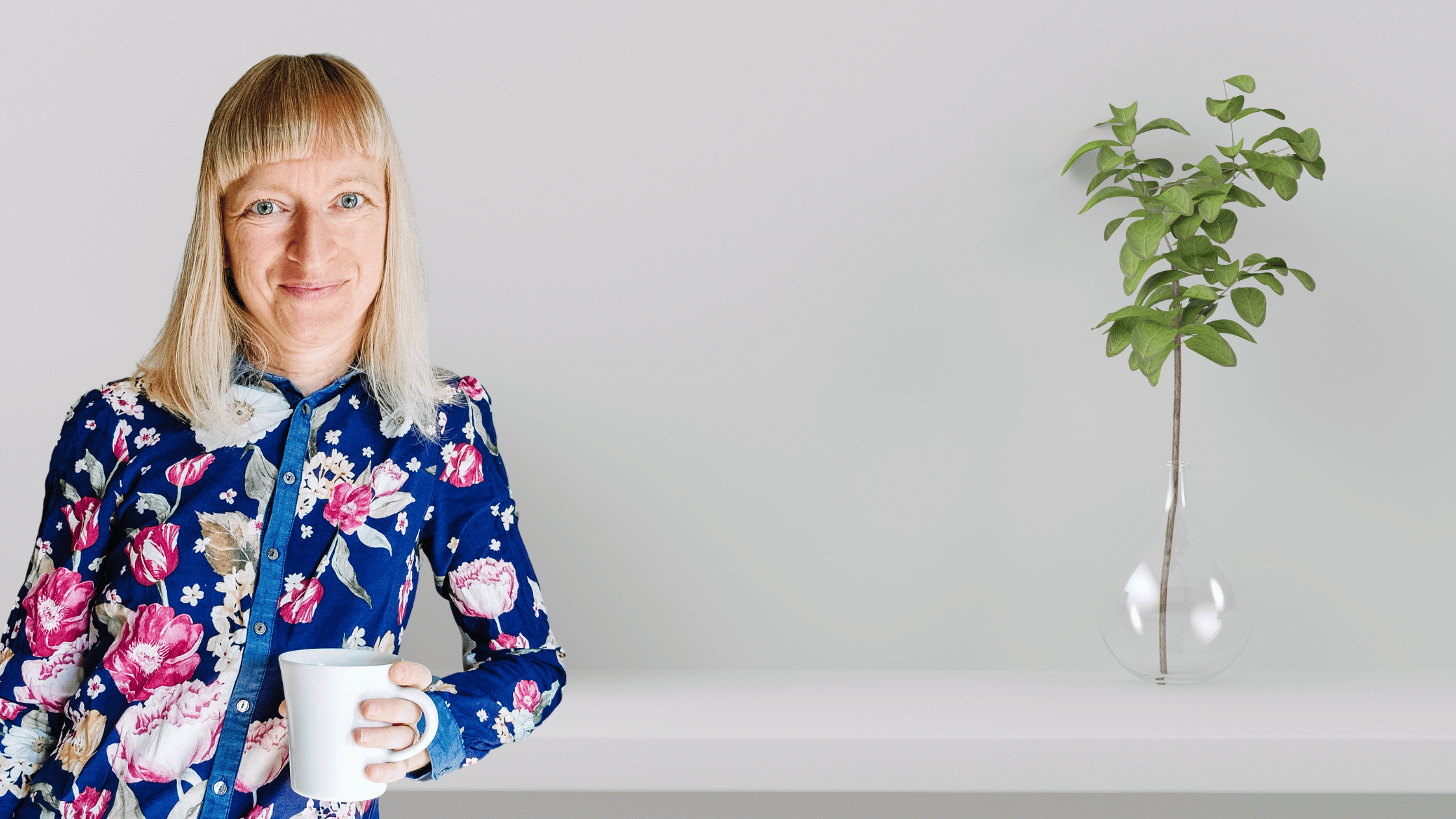 marlis schorcht steht mit kaffeetasse an einer theke lächelt rechts eine vase mit grünem zweig